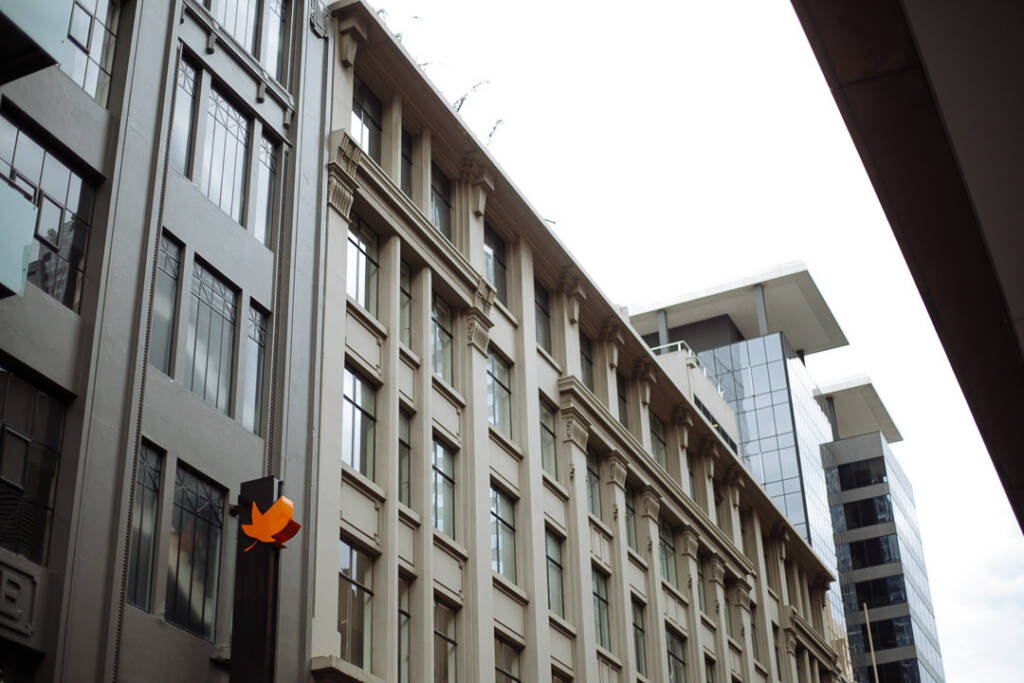 Looking up at a grey rendered facade with tall windows, an orange maple leaf sign on a post.