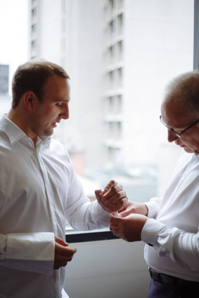 A grey haired man in glasses threads the groom's cufflink at the tower window, city buildings behind.