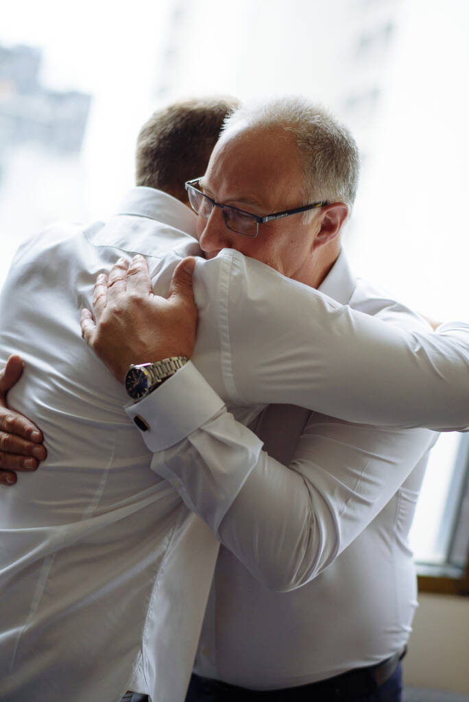 A long hug between the groom and the grey haired man in glasses at the bright window.