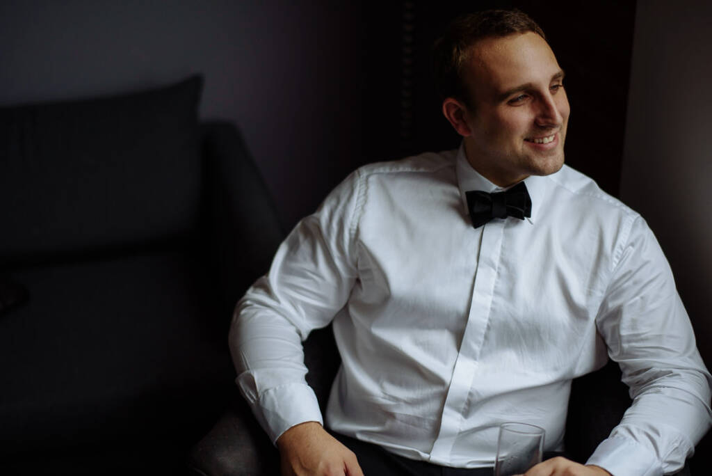 The groom smiles toward the window light from the armchair, glass at the ready.