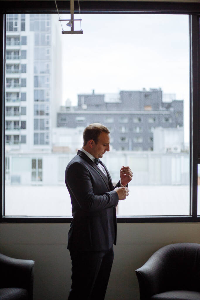 Silhouetted at the hotel glass, working the cufflink home