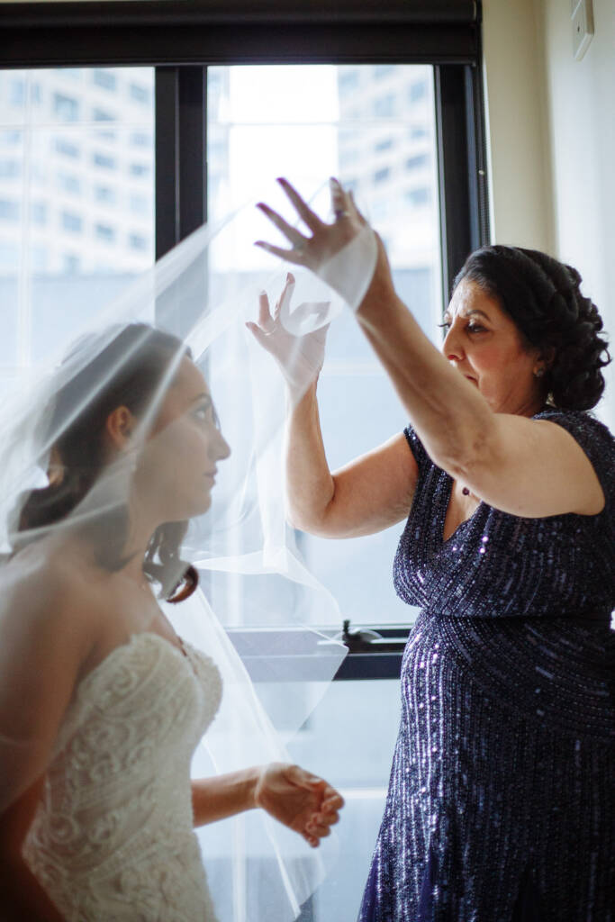 Two careful hands float the blusher over the bride's face at the window.