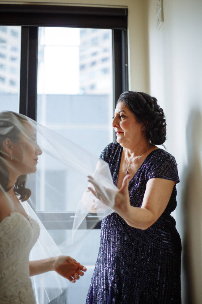 The woman in the sequinned navy gown frees the veil edge beside the bride in profile.