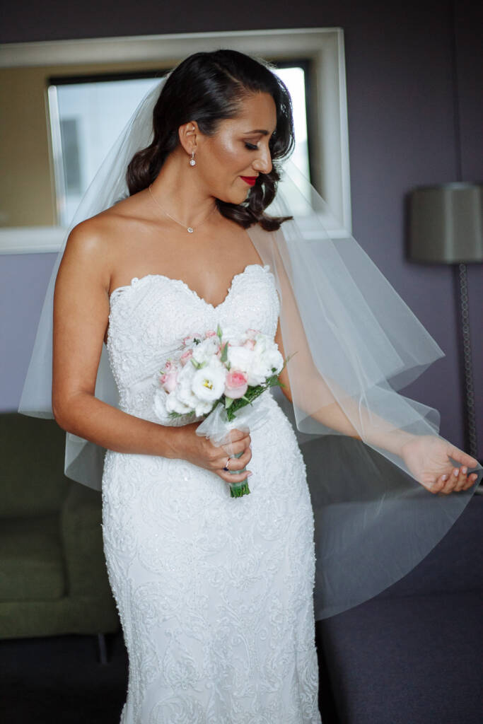 The bride smooths the veil back to admire the posy of pink roses and white lisianthus.