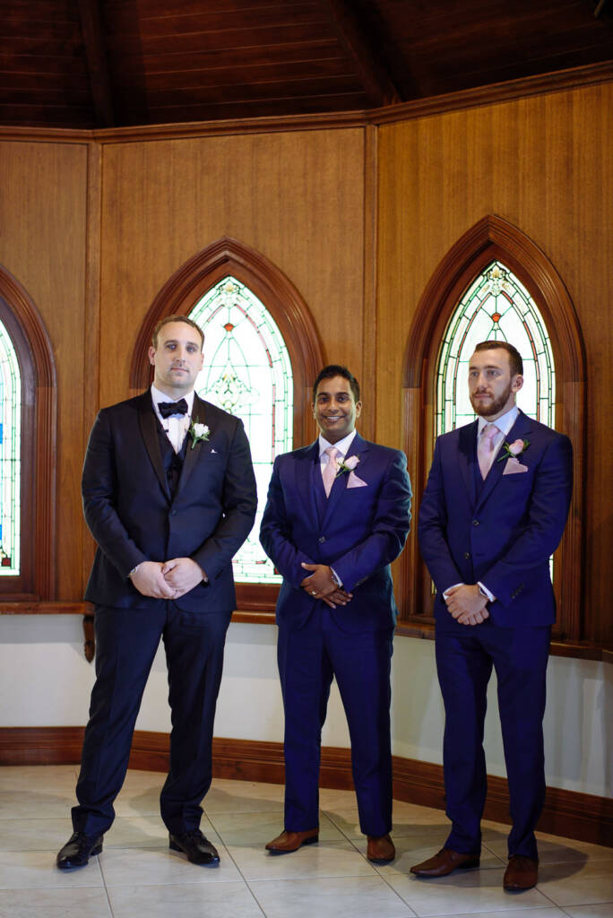 Groom and groomsmen wait square-shouldered before the stained glass