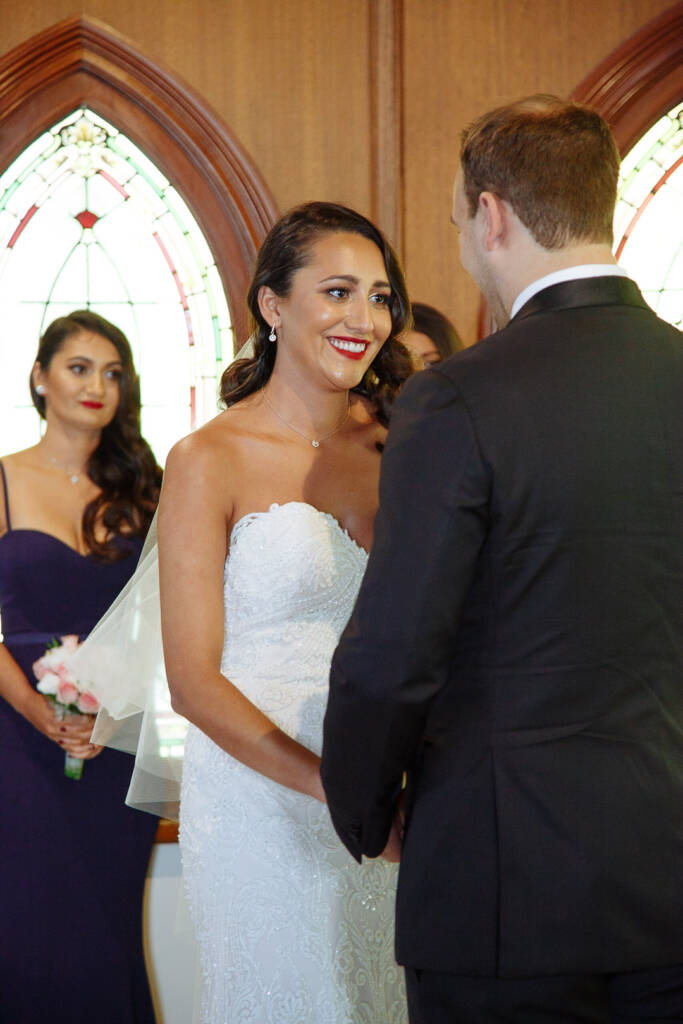 The bride's red lipped smile answers the groom in front of the arched stained glass.
