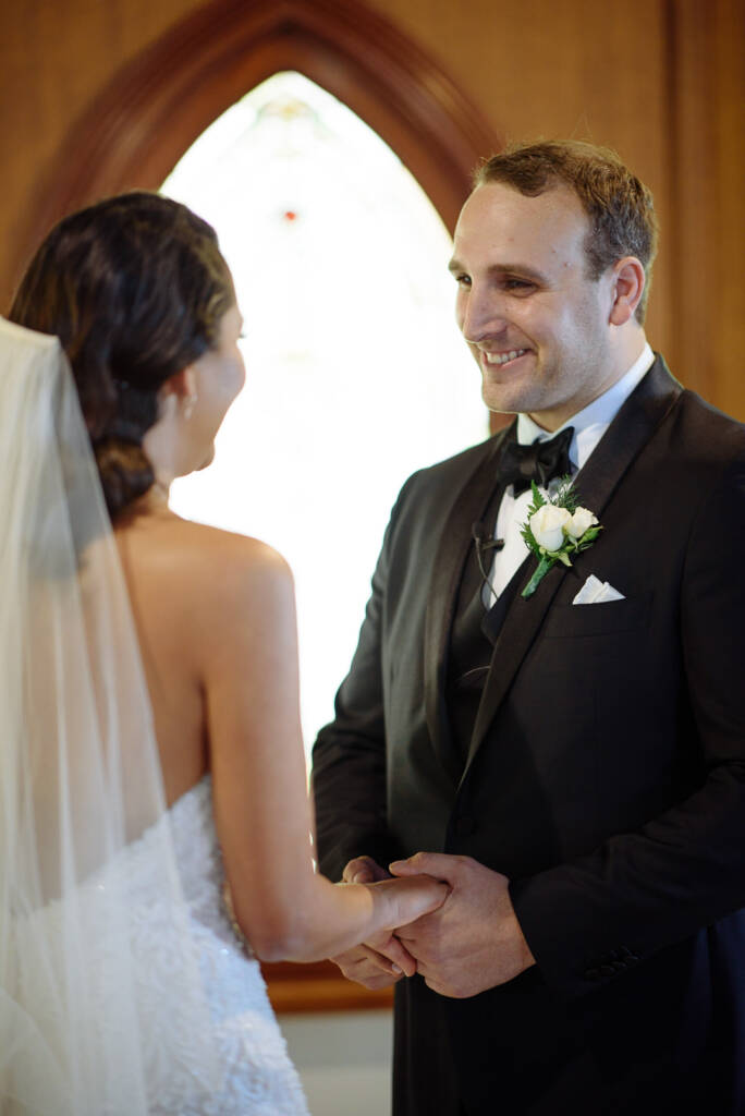 The groom's grin breaks over their joined hands at the altar, white rose on his lapel.