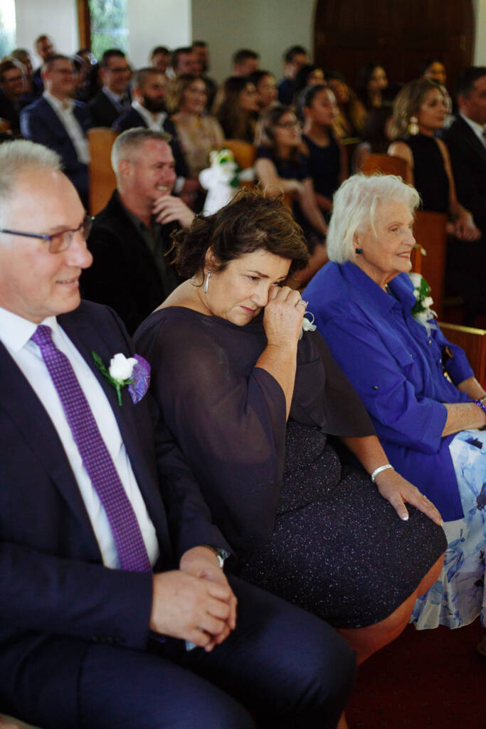 In the pew, a woman in navy chiffon wipes a tear beside a man in glasses.