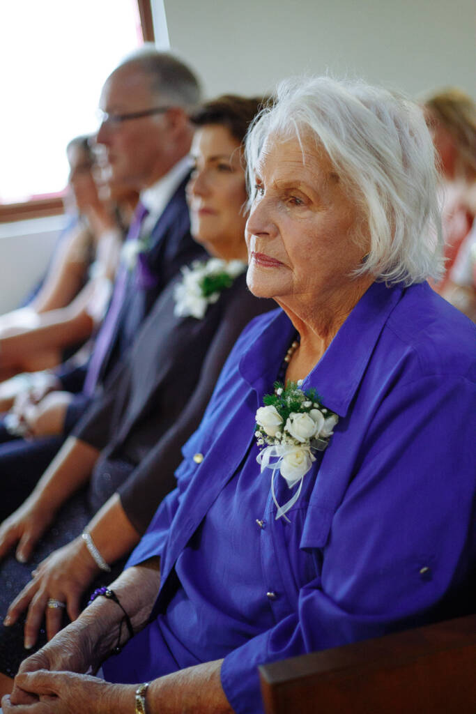 A white haired woman in a royal blue jacket watches from the pew with her white rose corsage.