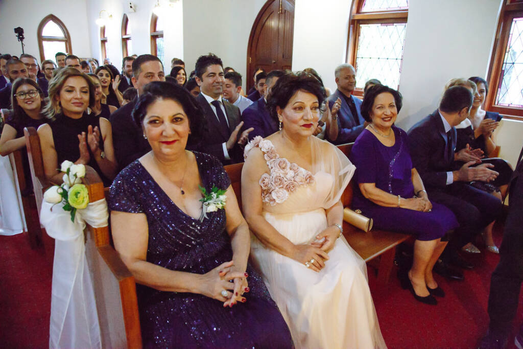Two women in blush and sequinned navy beam from the front bench as guests applaud behind them.