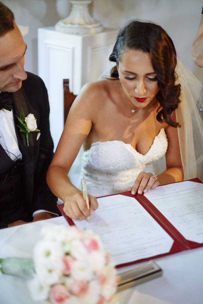 Head bowed, the bride signs the register with the beaded pen as the groom watches.