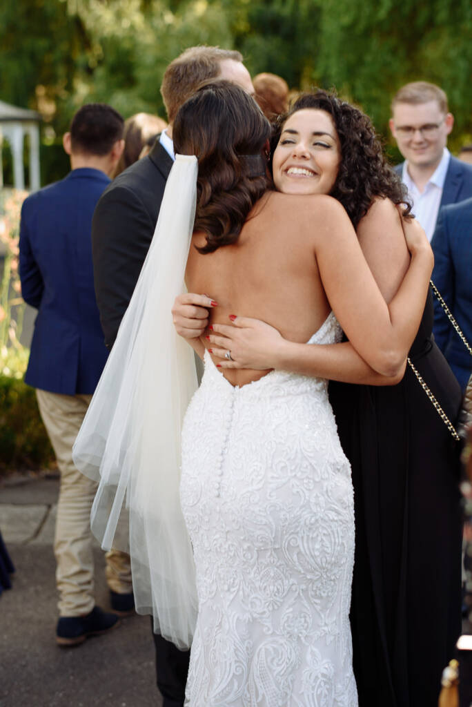 A curly haired woman hugs the bride tight, the bride's ring hand bright against her bare back.
