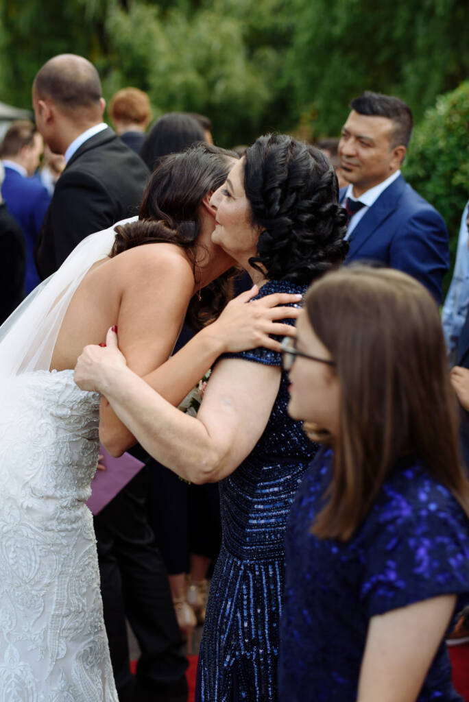 A woman with braided hair in sequinned blue folds the bride into another embrace outside the chapel.