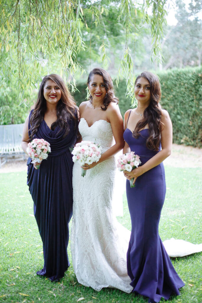 Willow fronds frame the bride between her two bridesmaids in navy, posies of pink roses.
