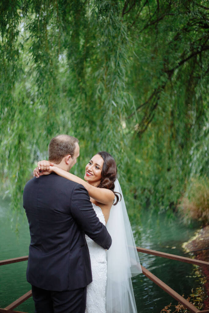 The bride looks back over the rusted rail, willows sweeping the lake behind the groom.