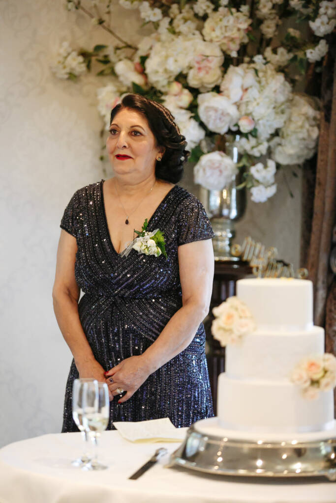 The woman in sequinned navy stands to speak beside the three tier cake and the blossom tree.