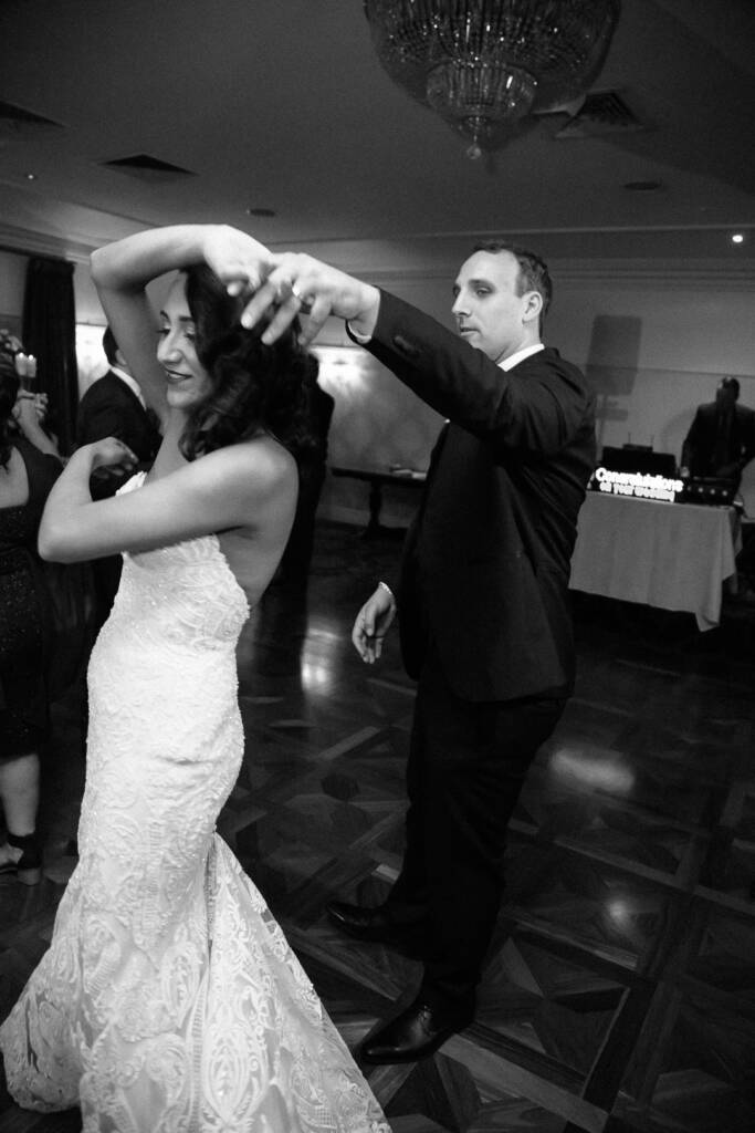 The groom sends the bride into a spin beneath the chandelier, the DJ booth lit behind.