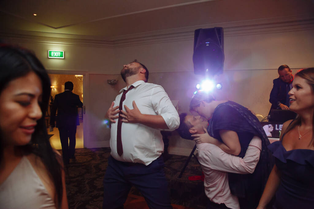 A guest in a loosened tie throws his head back as a dip becomes a kiss behind him.