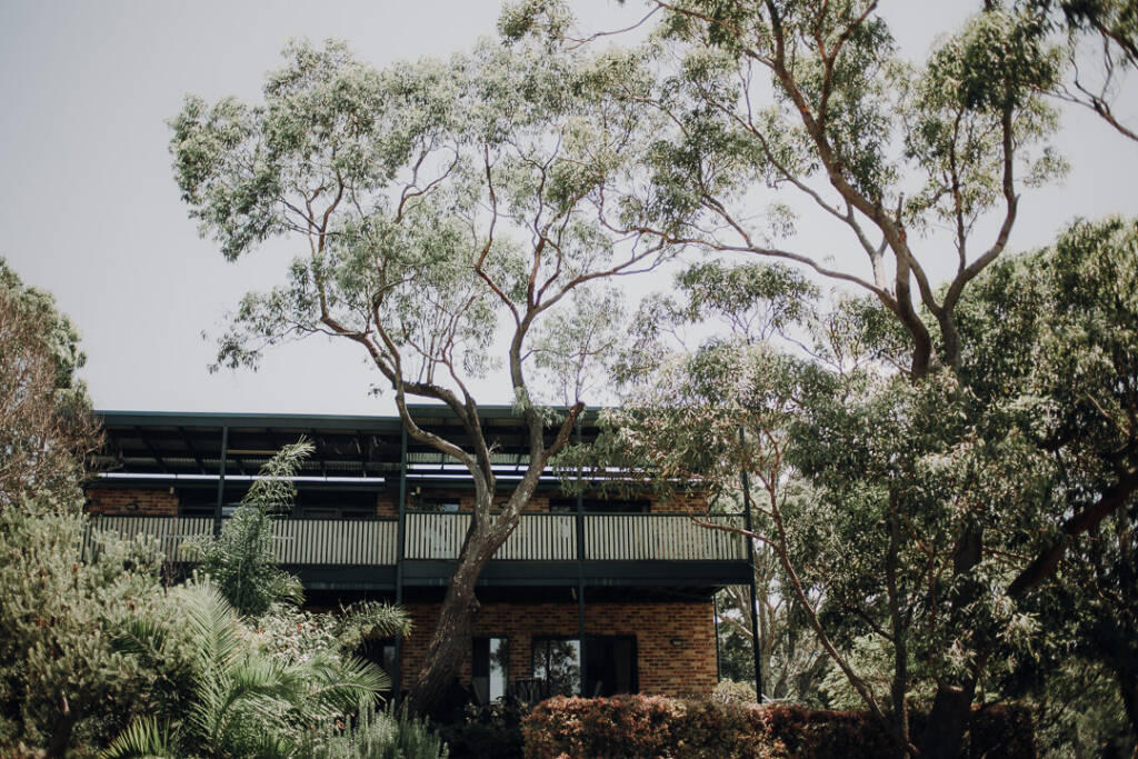 A two-storey brick home with a covered balcony stands behind trees in Allambie Heights, NSW.