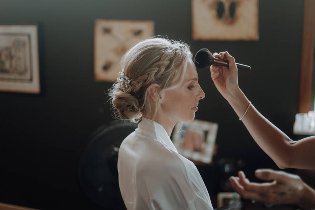 A makeup brush touches Emma’s forehead above her braided bridal hairstyle.