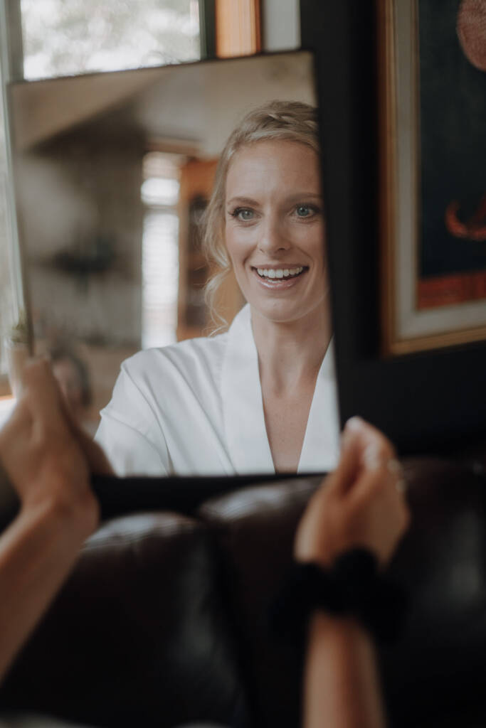 Emma smiles at her reflection in a handheld mirror during bridal preparation.