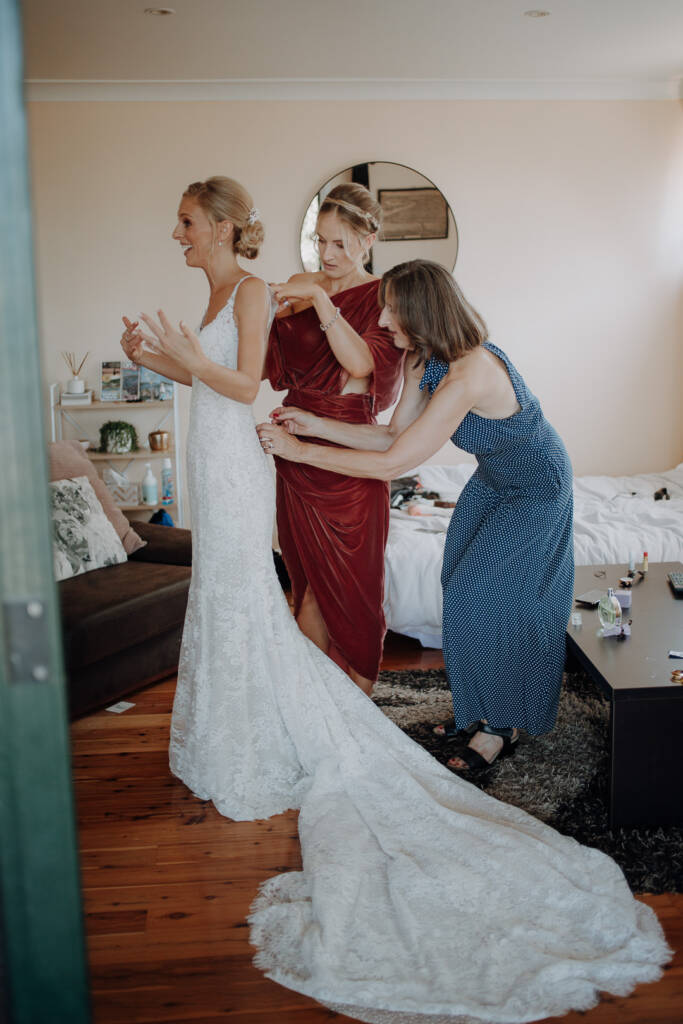 Two guests help Emma with her gown as its train spreads across a bedroom floor at a private home in Allambie Heights.