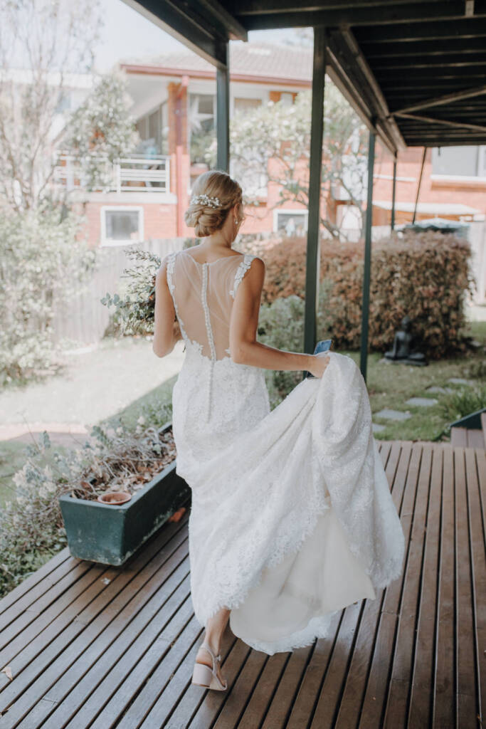 Emma lifts her lace train while walking in block-heeled shoes along the timber deck of a private home in Allambie Heights.