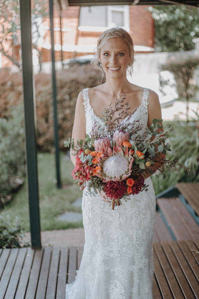Emma smiles towards the camera with her pink protea and orange-flower bouquet.