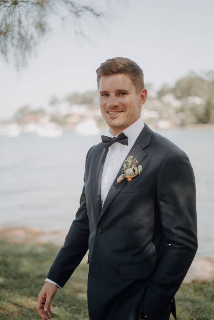Sean poses beside Pittwater near BYRA in his dark wedding suit and boutonniere.