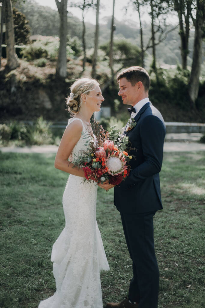 Emma and Sean face each other with her protea bouquet between them near Bayview Yacht Club.