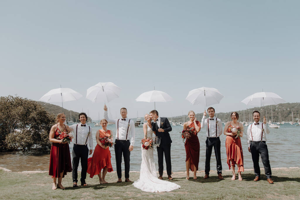 The wedding party pose with white umbrellas beside Pittwater near Bayview Yacht Racing Association.