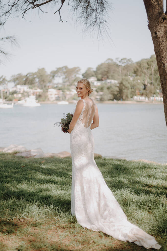 Emma looks back beneath a tree near Bayview Yacht Club, holding her bouquet beside her train.