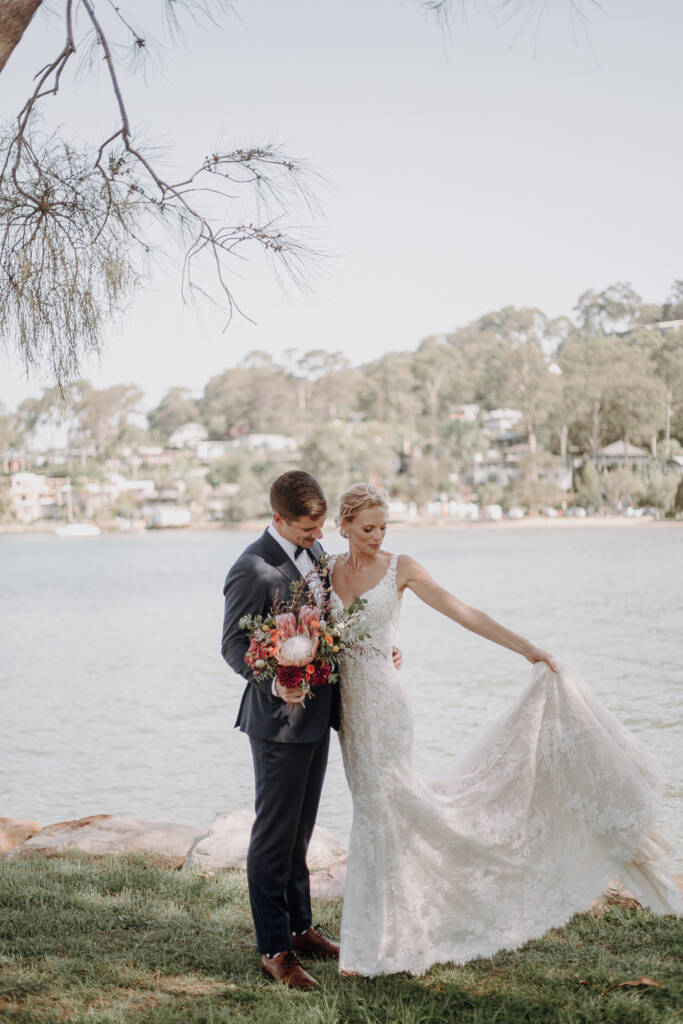 Sean holds the bouquet while Emma gathers her lace train beside Pittwater near BYRA.