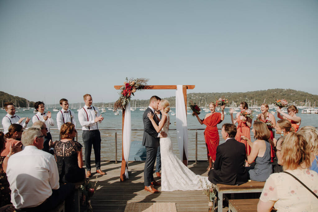 Emma and Sean kiss beneath BYRA’s wedding arbour as their attendants applaud.