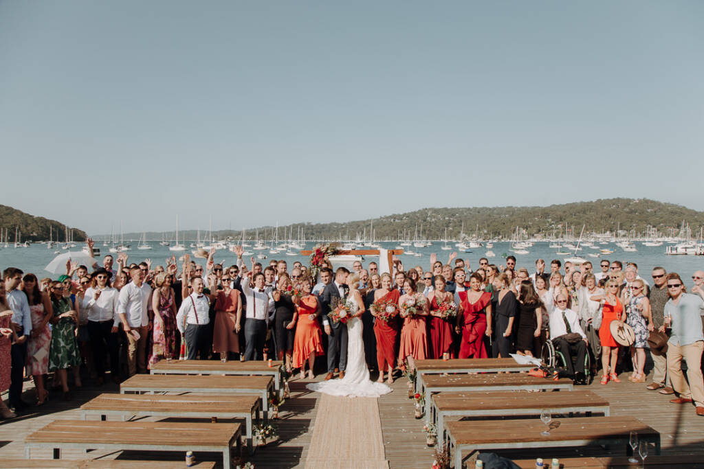 Guests gather around the couple for a group wedding photo on Bayview Yacht Club’s deck above Pittwater.