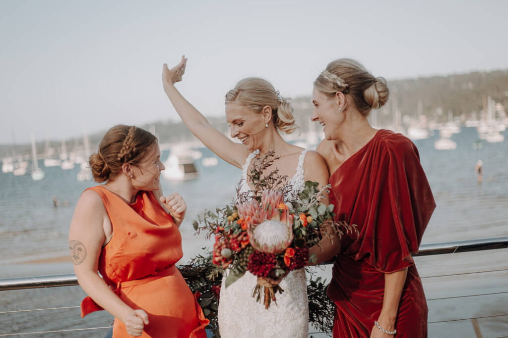 Emma raises an arm and smiles between two attendants beside Bayview Yacht Club’s deck railing.