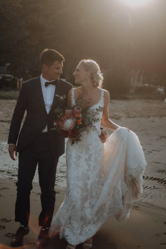 Emma lifts her train as she walks beside Sean in warm backlight near BYRA on the Northern Beaches.