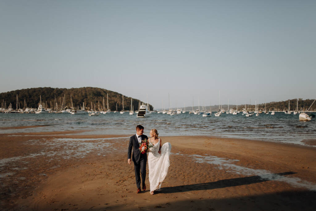 Emma and Sean walk across the sandy beach near Bayview Yacht Racing Association, with moored yachts behind them.