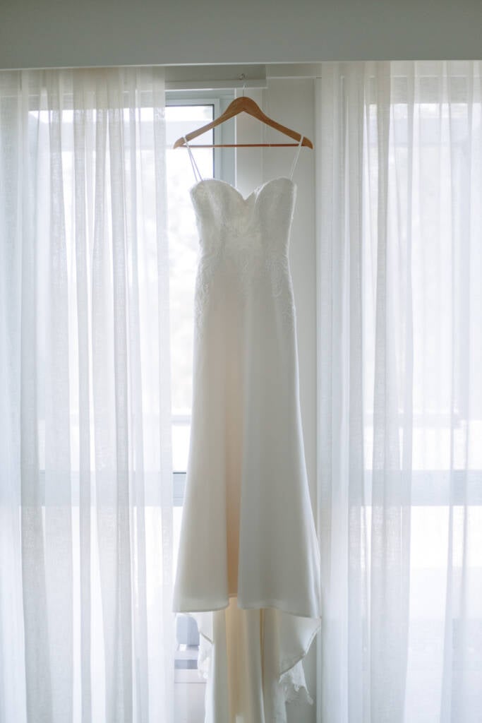 Becky's strapless wedding gown hangs against sheer curtains before the Greek islander wedding.