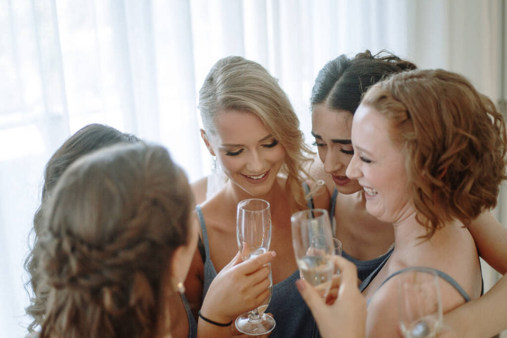 The bridal party laugh together over champagne during candid wedding preparations.