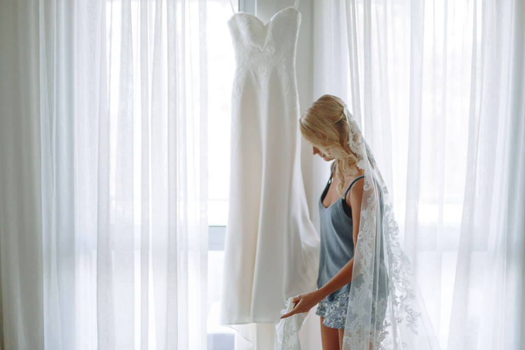 Becky stands in her lace veil beside her hanging wedding gown during bridal preparation.