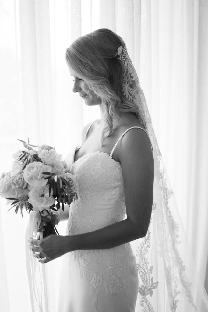 Black and white bridal portrait of Becky holding her blush rose bouquet beside a window.