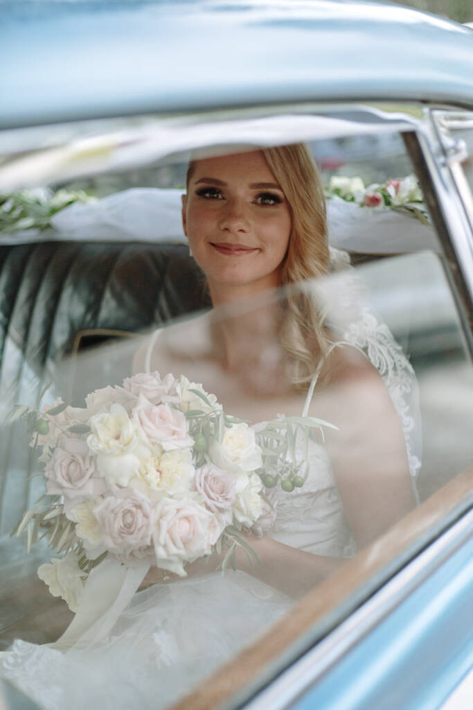 Becky smiles through the window of a powder-blue vintage wedding car while holding her bouquet.