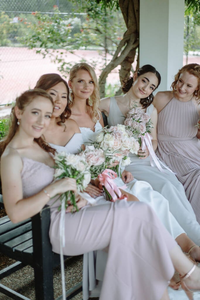 Becky and her bridesmaids sit with pastel bouquets on a garden bench at Old Parliament House Gardens.