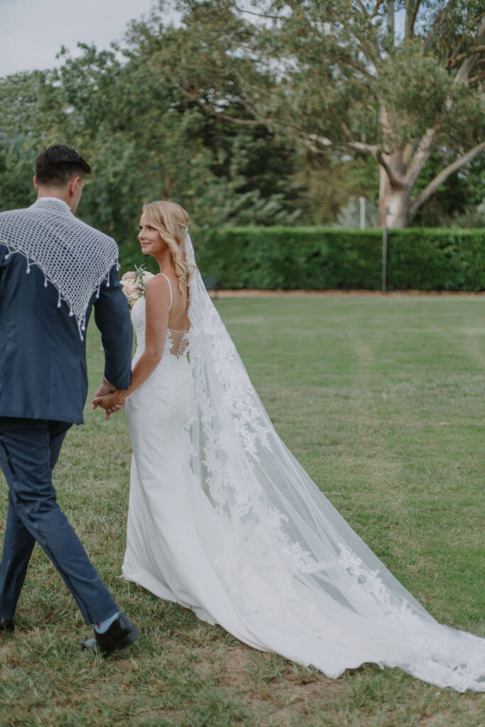 Becky's long appliquéd veil trails across the lawn as she walks with George at Senate Gardens Canberra.