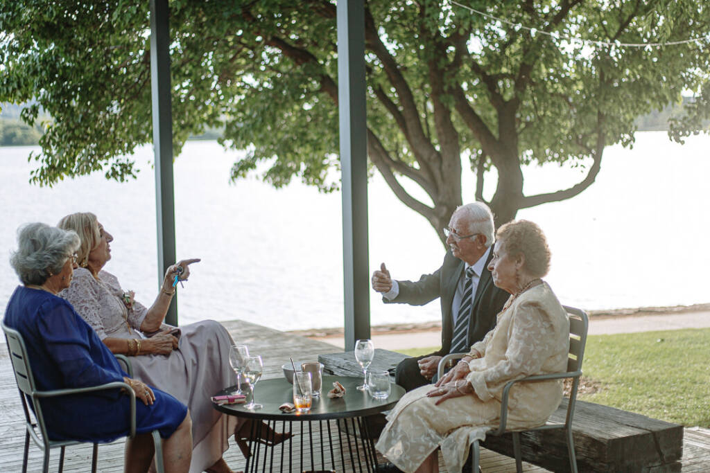 Wedding guests talk at an outdoor table beneath a tree at Boat House by the Lake.