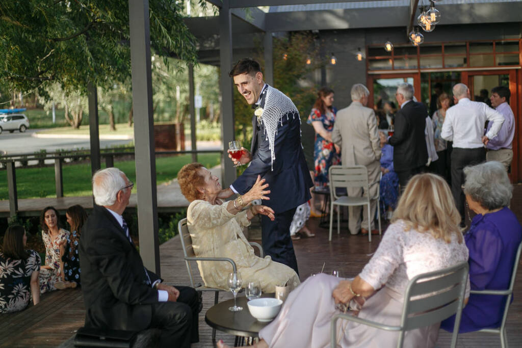 George greets a seated wedding guest on the waterfront terrace at The Boat House Canberra.