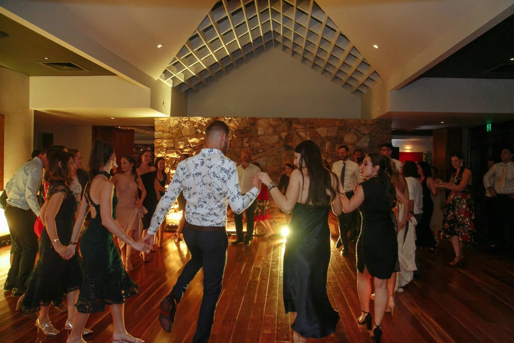 Guests form a lively circle dance beneath the timber ceiling at The Boat House Barton during the Greek islander reception.