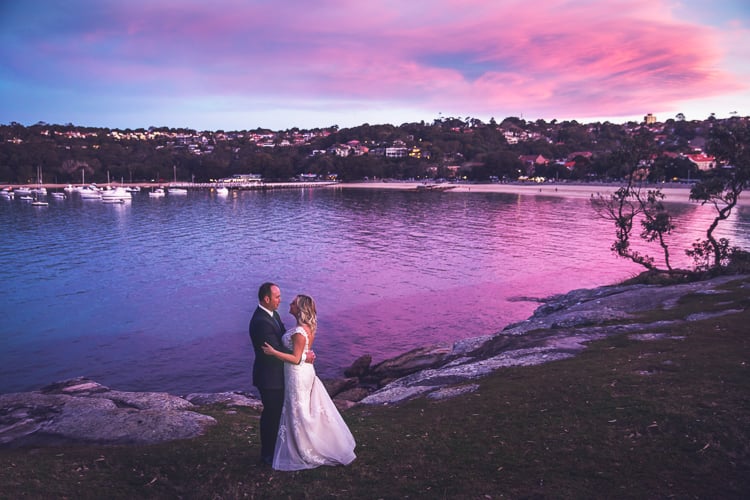 Sabine and Arthur embrace on waterfront rocks near Bathers' beneath vivid sunset clouds