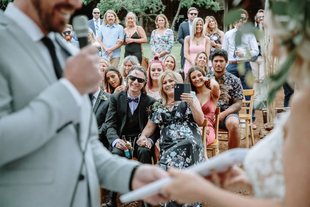 Seated guests smile and hold phones during the outdoor wedding ceremony.